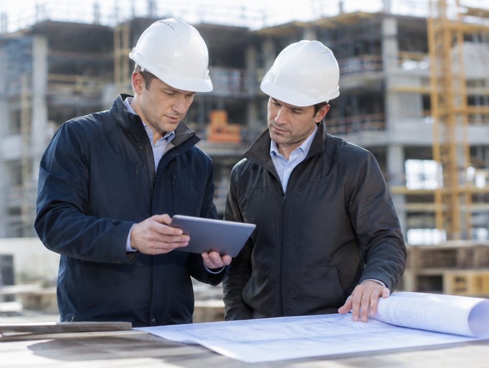 Two project managers in hard hats reviewing a schedule on a tablet and drawings on site